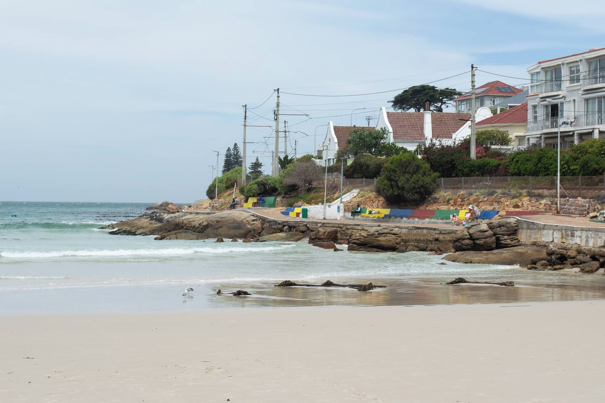 Uma praia costeira com afloramentos rochosos, um calçadão e edifícios residenciais ao fundo.