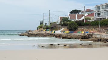 Uma praia costeira com afloramentos rochosos, um calçadão e edifícios residenciais ao fundo.