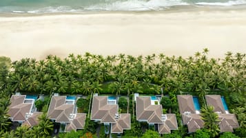 An aerial view of a beachfront resort with multiple villas, a pool area, and dense palm trees.