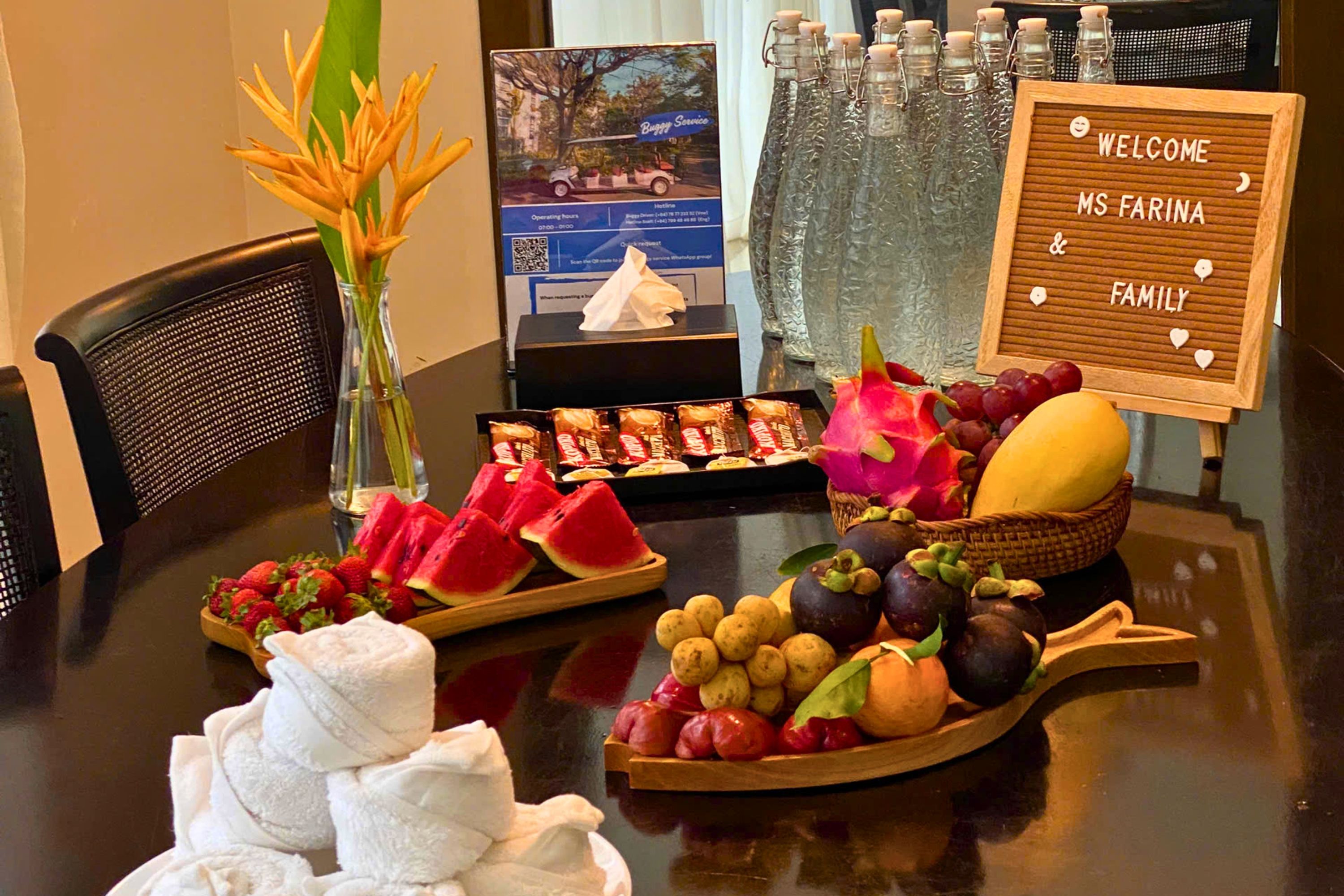A table with a welcome sign, snacks, fruits, and a vase with flowers.