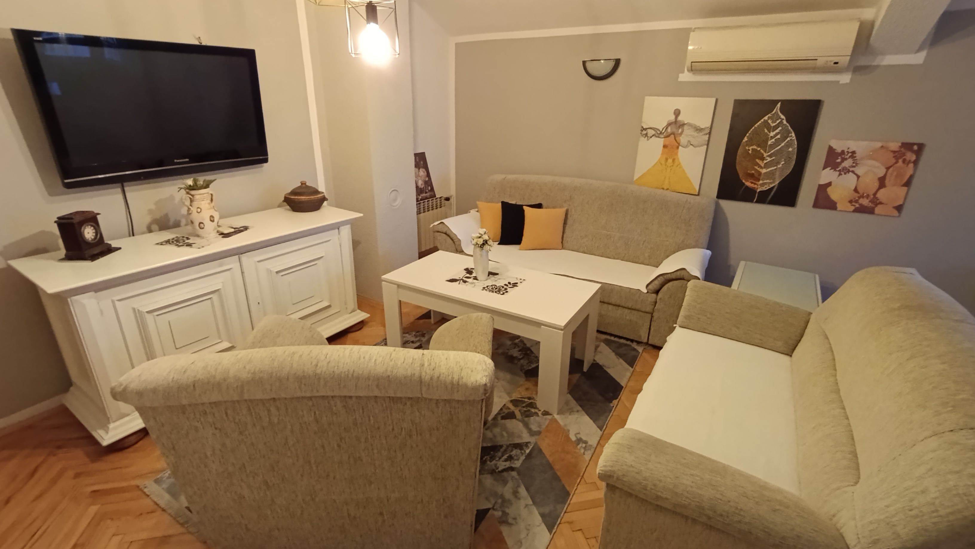 A living room with a flat-screen TV, a white table, a beige sofa, and a patterned rug.