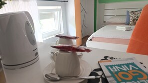 Una habitación de hotel con cama, una mesita de noche con tetera, un plato, una cuchara y una revista sobre la mesa.