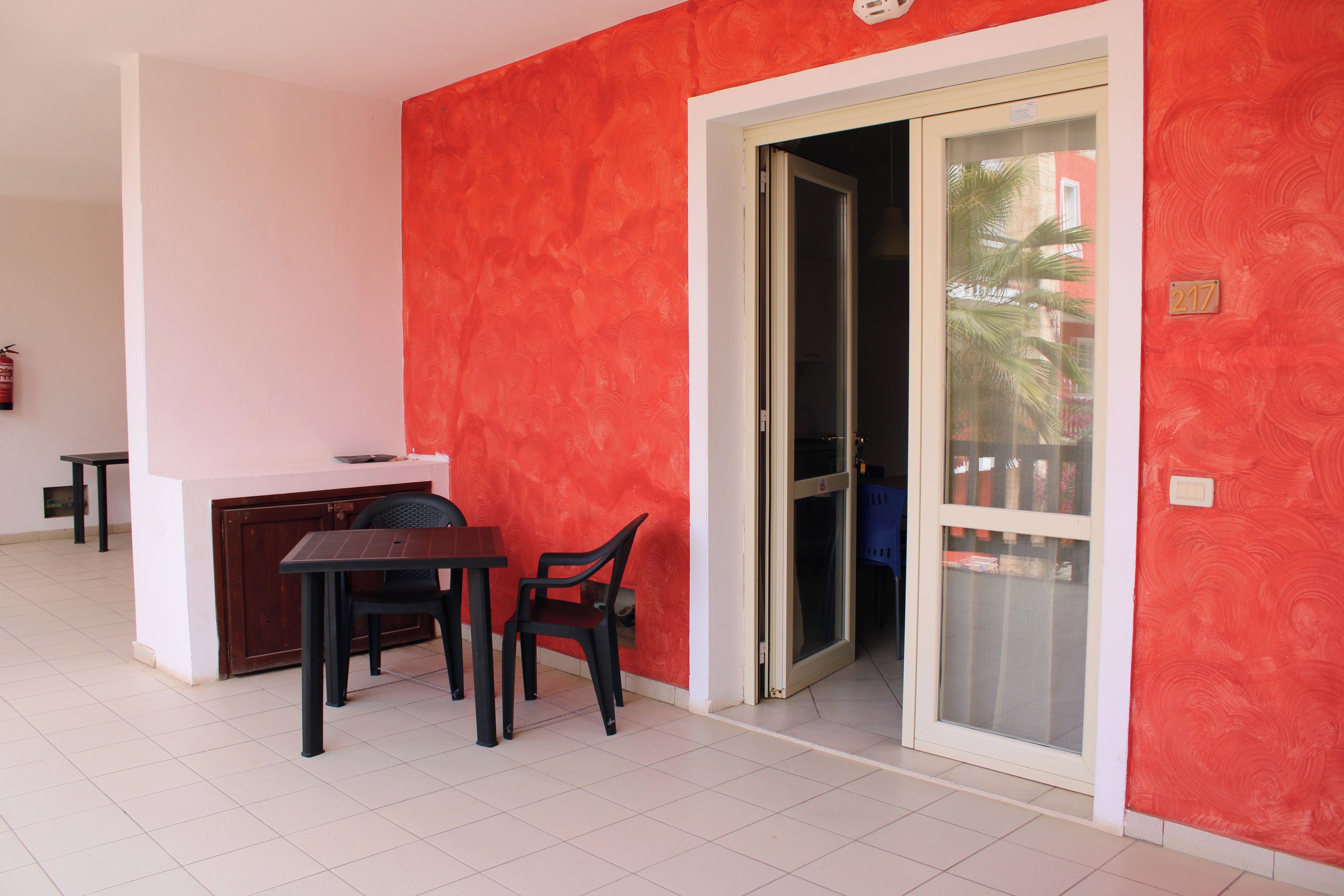 Una habitación con una pared roja, puerta corredera de vidrio, una mesita con dos sillas y un techo blanco.