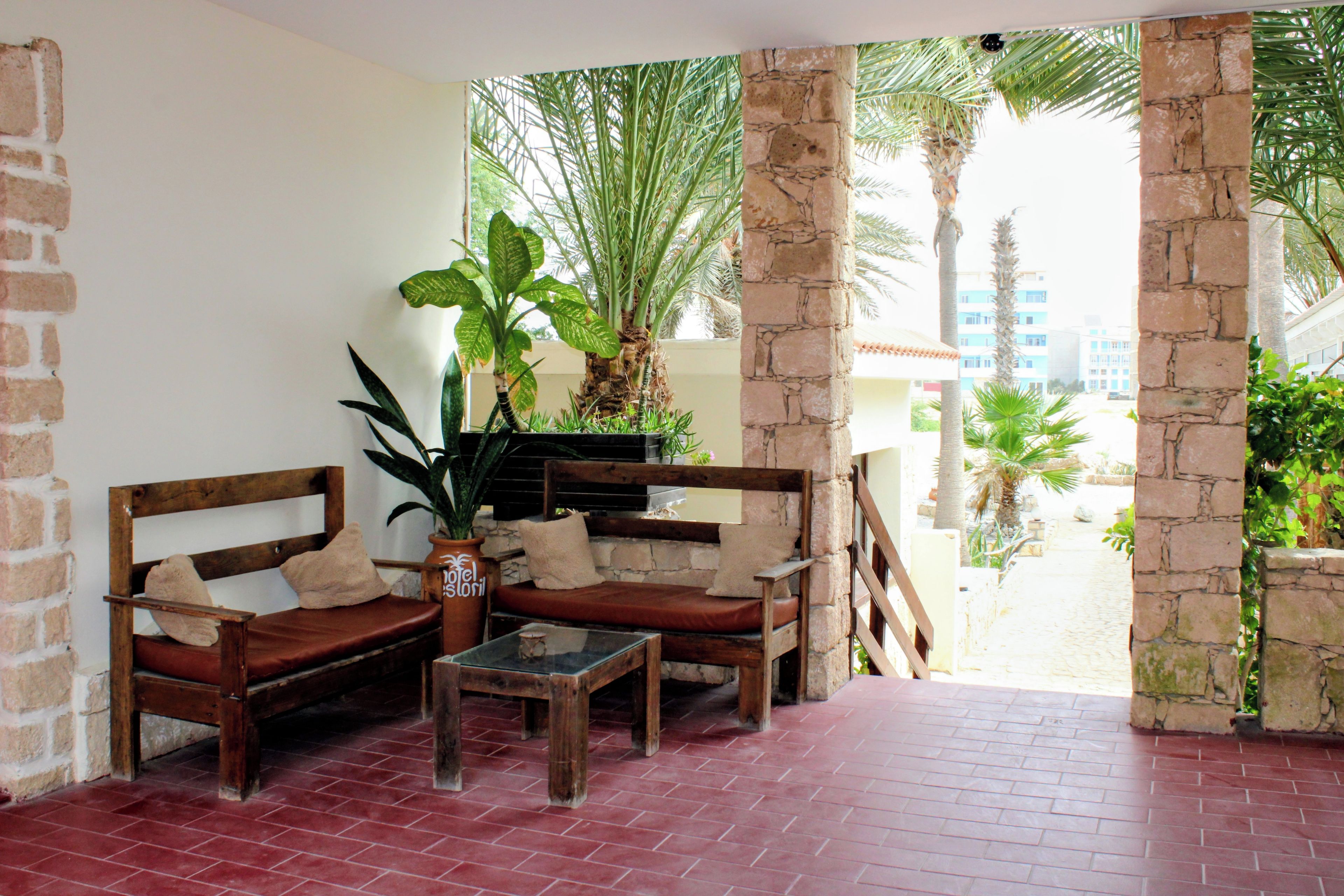 A room with a brick floor, wooden furniture, and a view of palm trees and buildings.