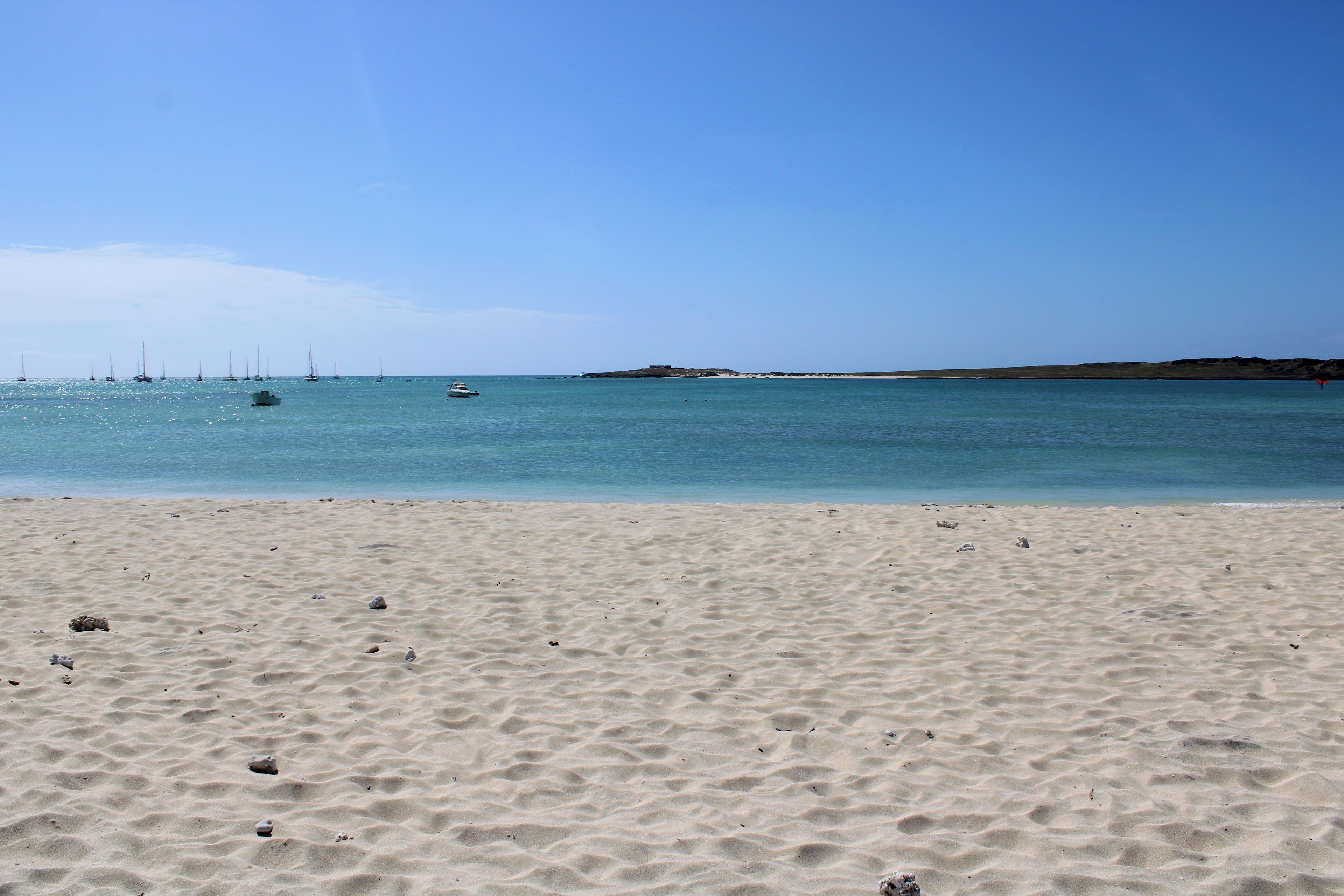 Une plage de sable fin, avec une eau calme et des bateaux visibles au loin.