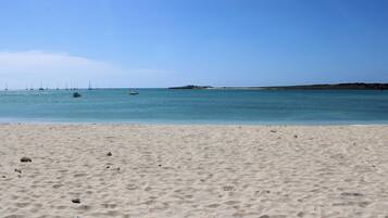 Une plage de sable fin, avec une eau calme et des bateaux visibles au loin.