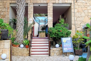 A stone building with a staircase leading to a door, surrounded by potted plants and a signboard.