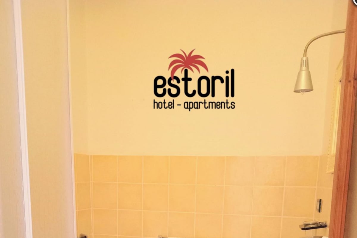 Un baño con inodoro, bidé y lavamanos, con pared de azulejos y el logotipo de 'estoril hotel - apartments'.