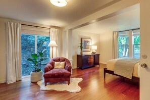 A spacious room with hardwood flooring, a plush armchair, a bed with a striped blanket, and a large window offering a view of greenery. - Updated Bungalow in The Dalles! Walk to Downtown! (The Dalles)