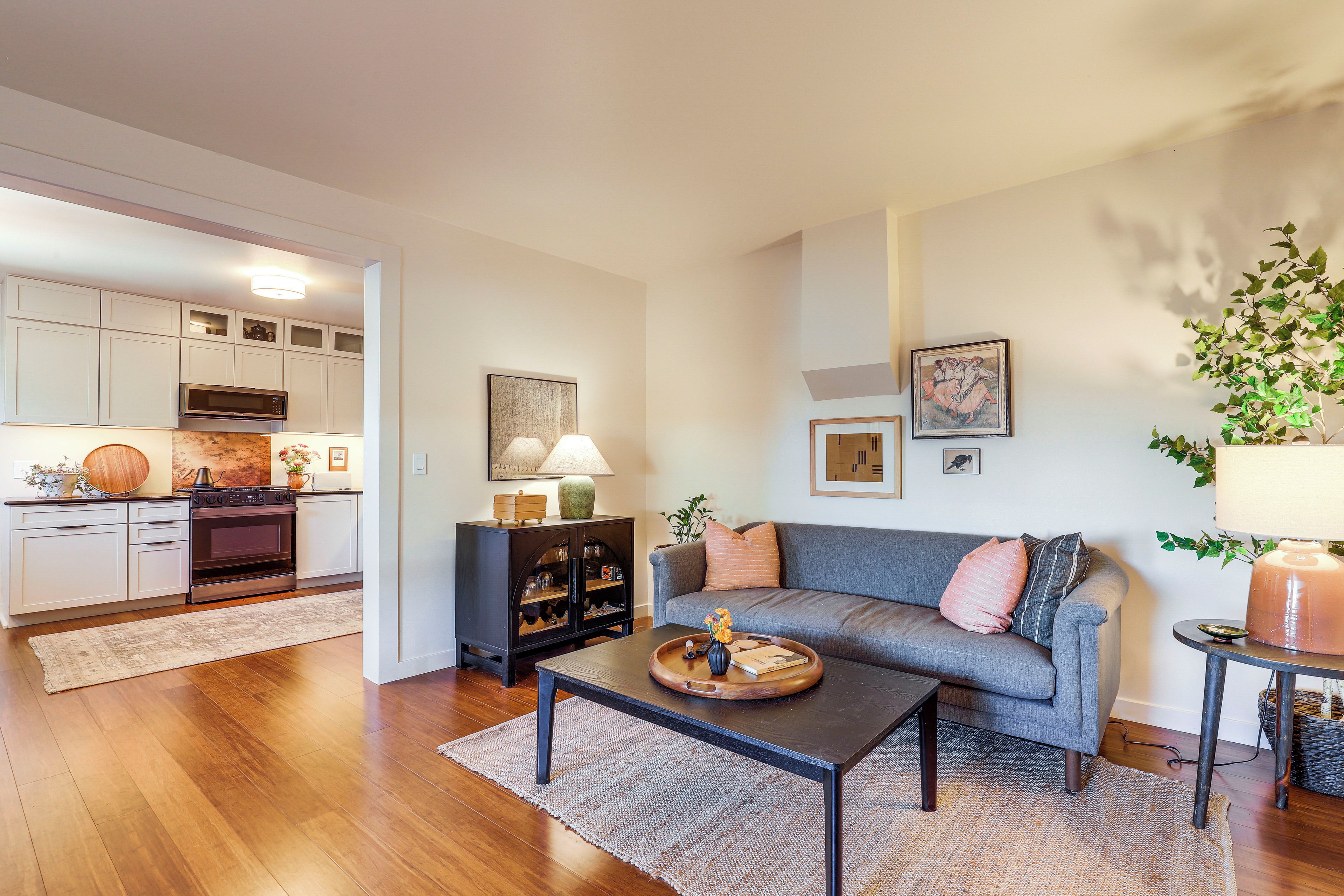 A modern living room with a grey sofa, coffee table, and a kitchen area with white cabinets and appliances.
