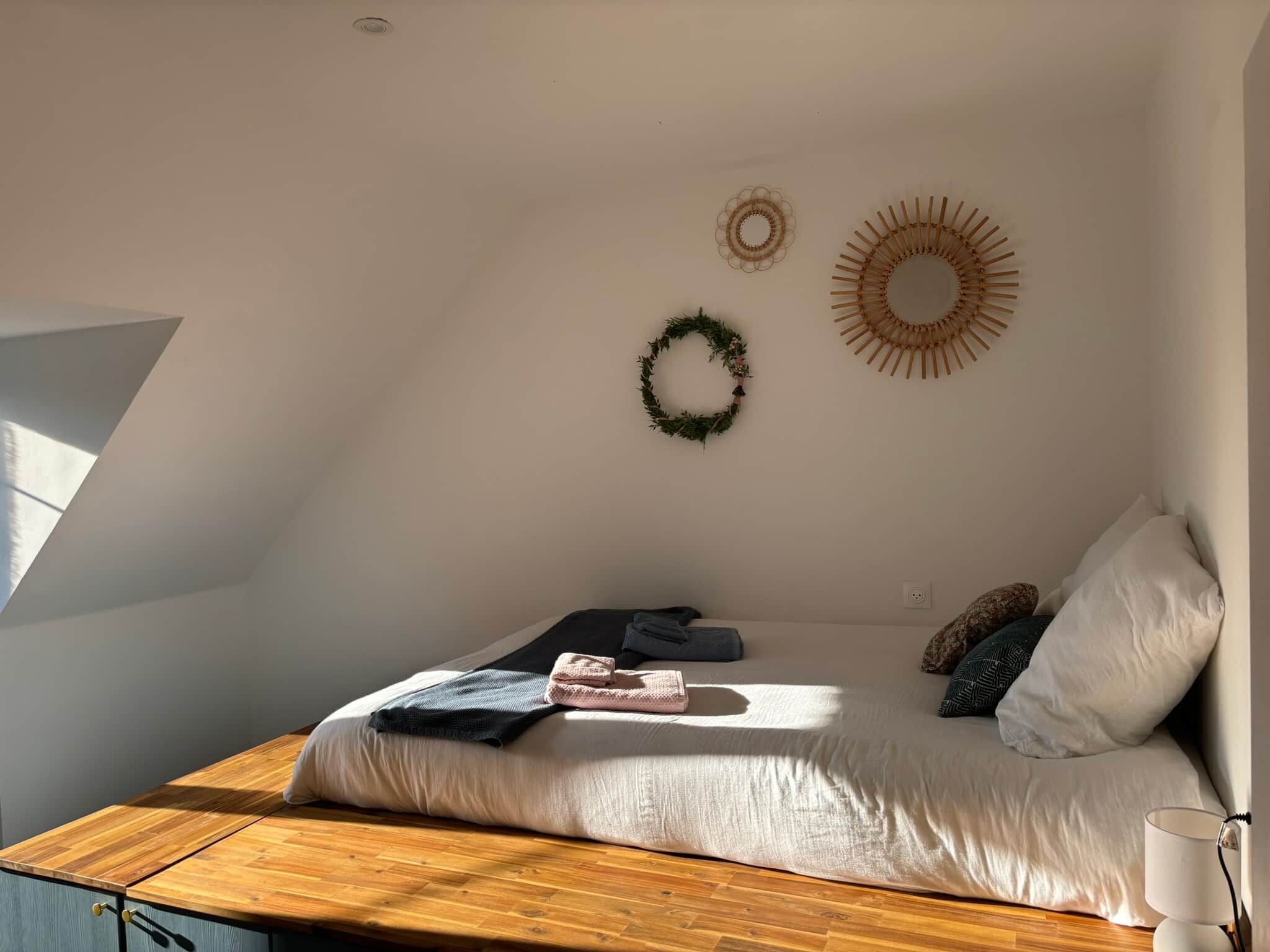 Une chambre avec un plafond en pente, un lit avec des oreillers et une couverture, une table en bois et des éléments de décoration murale, notamment une guirlande et un motif en forme de soleil.
