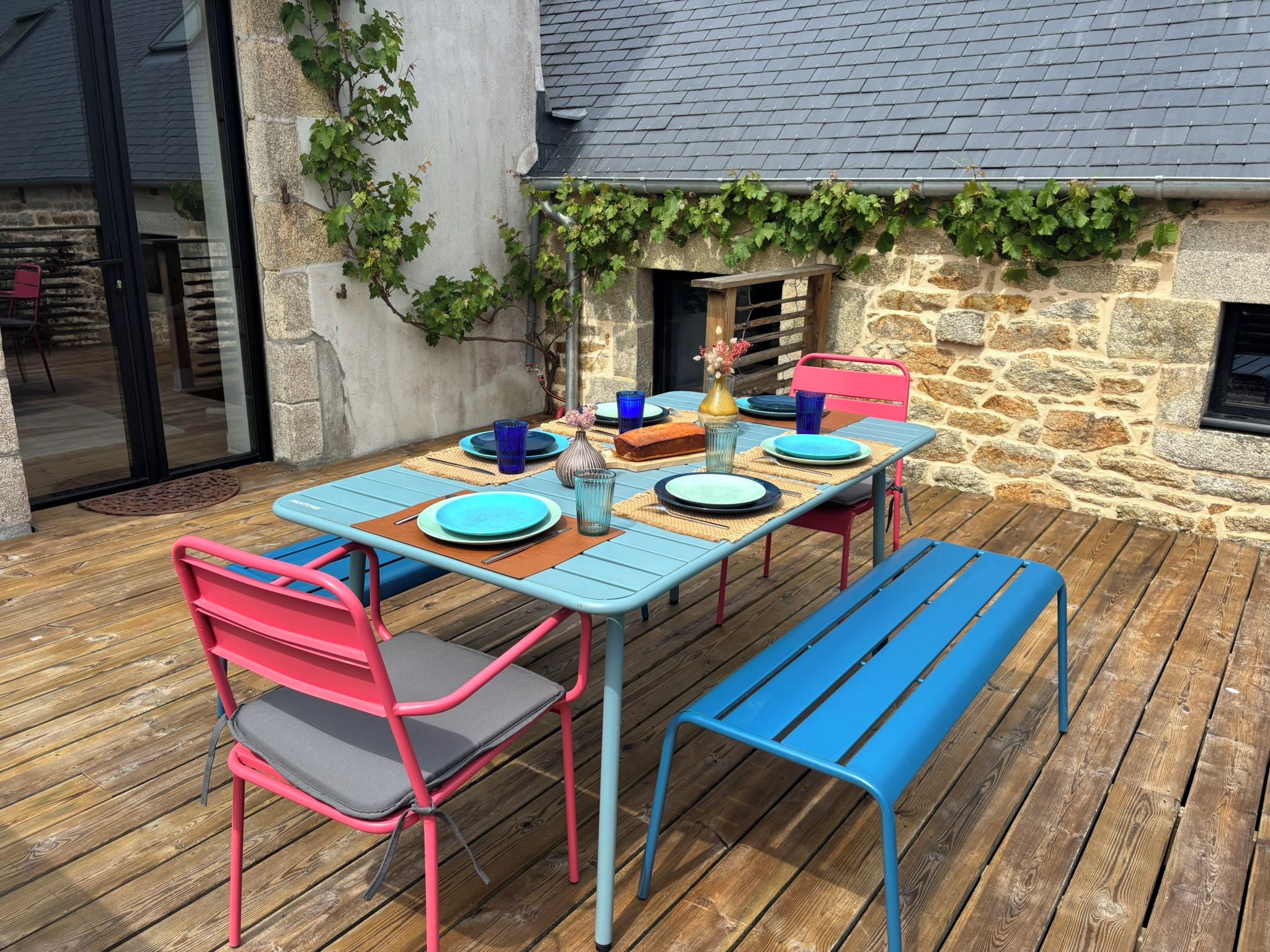 Un espace extérieur aménagé pour les repas, avec un mur en pierre, une terrasse en bois et une table dressée avec des assiettes et des chaises bleues.