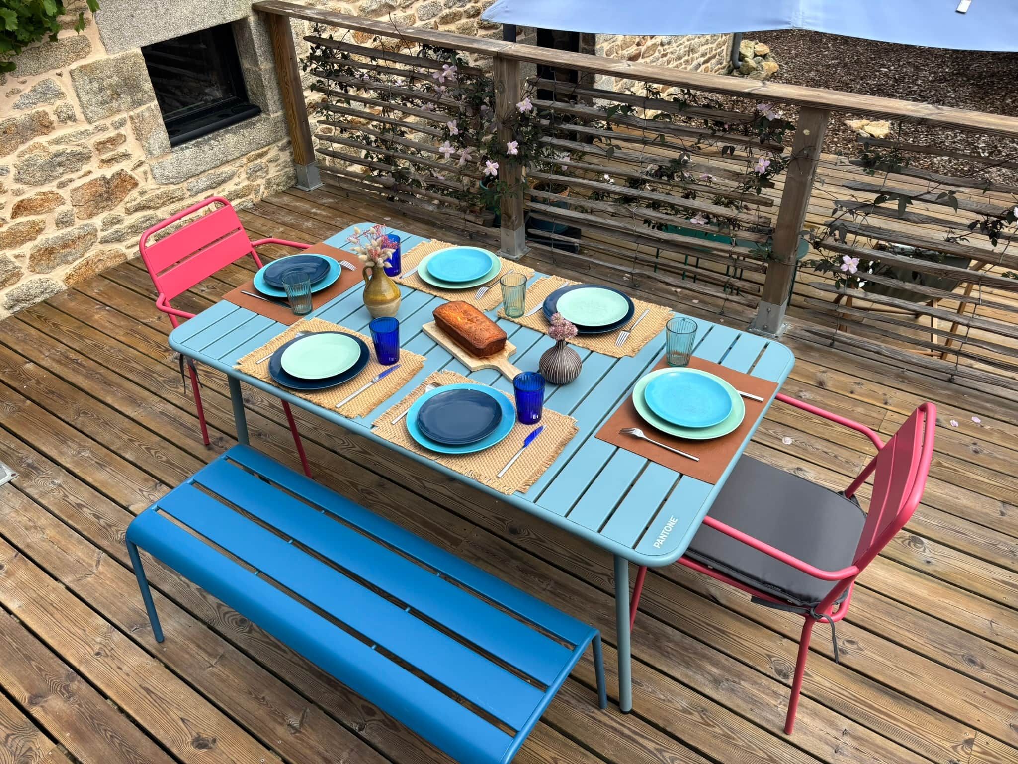 A wooden deck with a table set for two, featuring blue and red chairs, a bench, and a stone wall in the background.