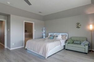 Un dormitorio con cama, mesita de noche, silla, estante en la pared y puerta que da a un baño.