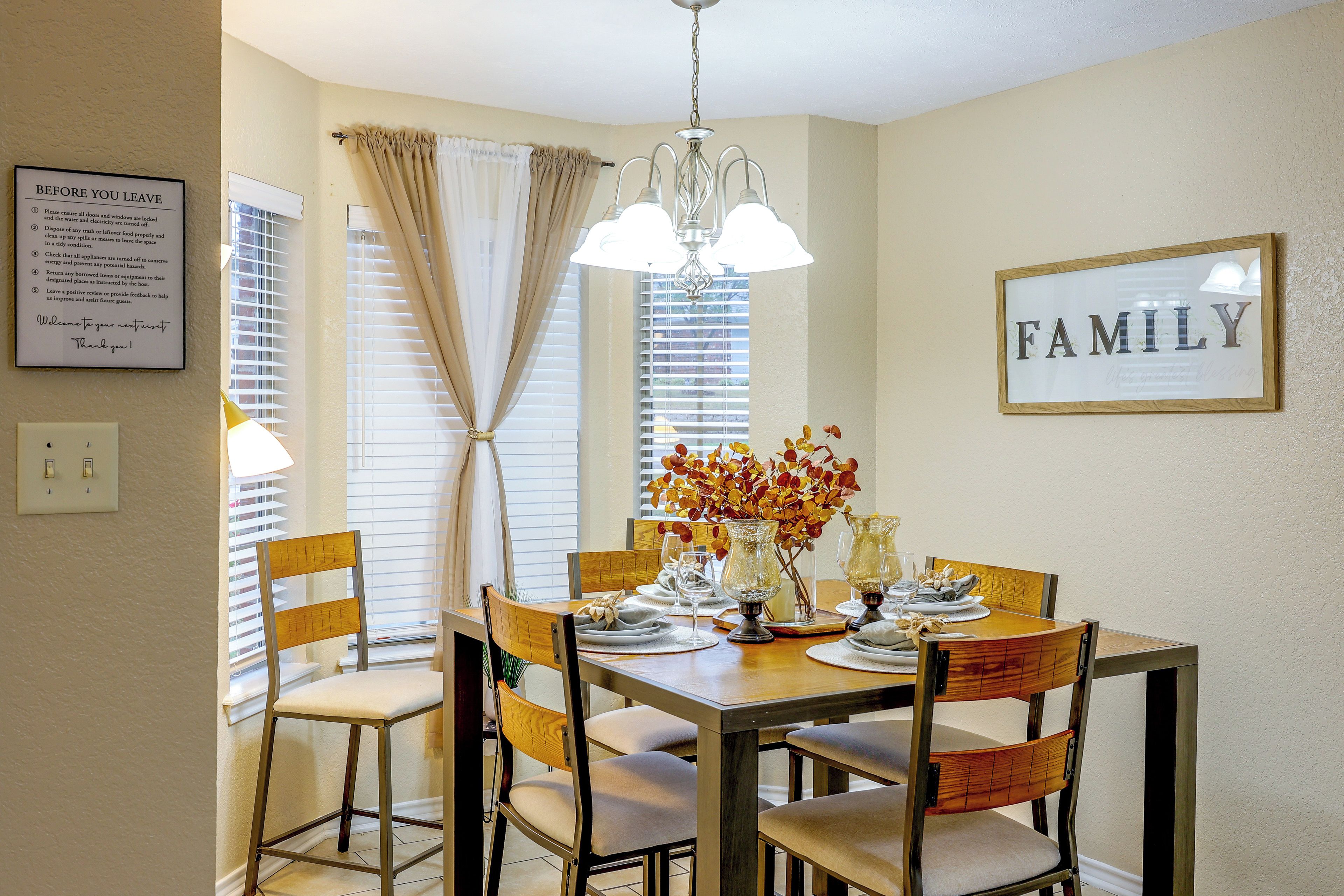 A dining area with a wooden table set for four, surrounded by chairs. A chandelier hangs from the ceiling, and a framed sign on the wall reads 'FAMILY.'