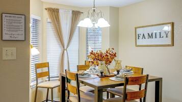 A dining area with a wooden table set for four, surrounded by chairs. A chandelier hangs from the ceiling, and a framed sign on the wall reads 'FAMILY.'