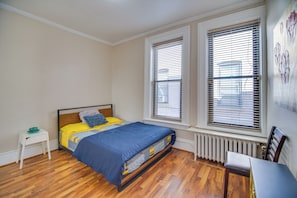 Un dormitorio con una cama, una mesita de noche, una silla y dos ventanas con persianas.