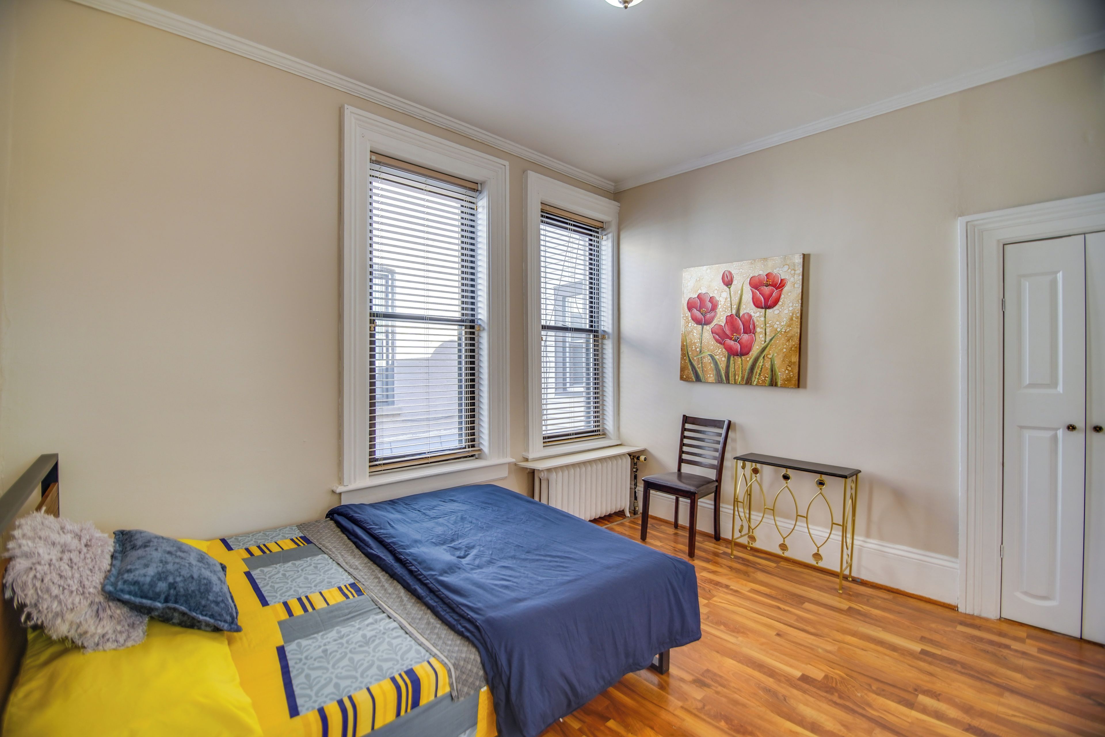 Un dormitorio con una cama, dos ventanas con persianas, una silla, un radiador y una puerta.