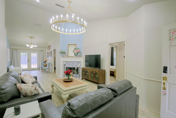 A living room with a fireplace, a television, and a chandelier. - The Iola / Chic Vintage Glam & Modern Comfort (Lubbock)