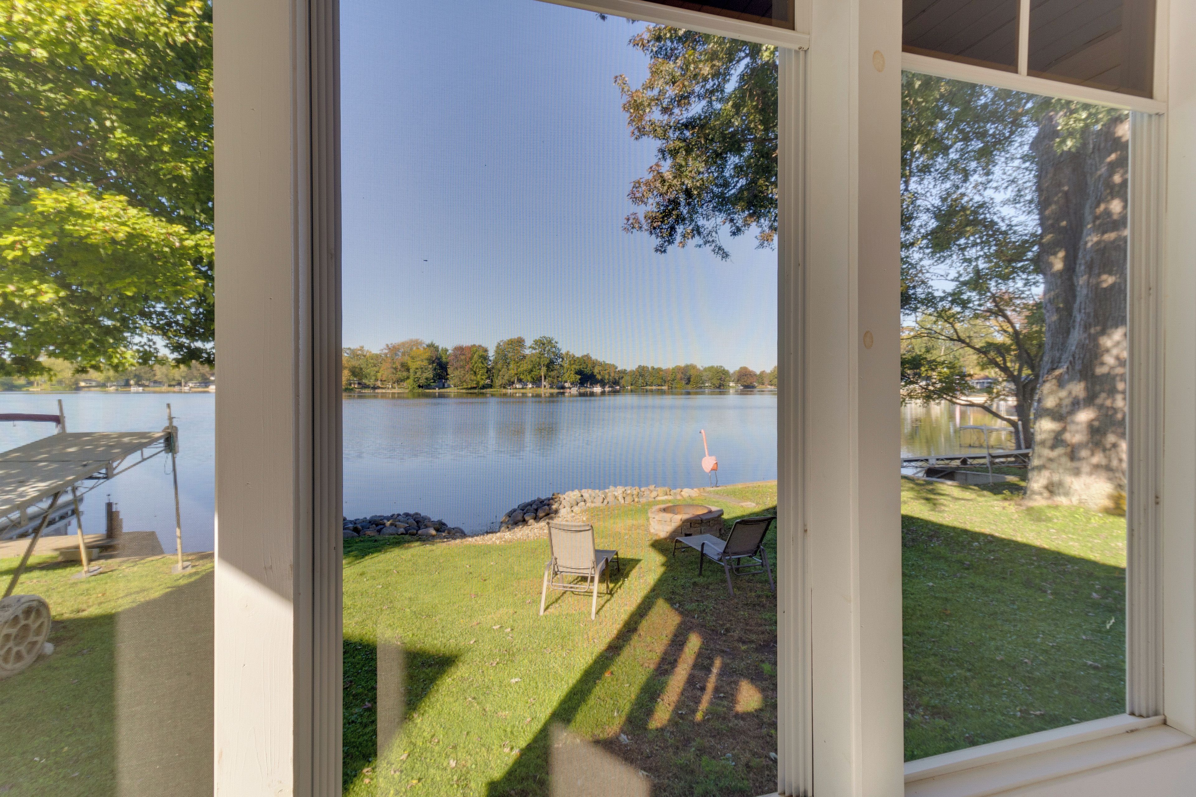 A view through a screened door of a lakeside property with a deck, chairs, and a flamingo statue.