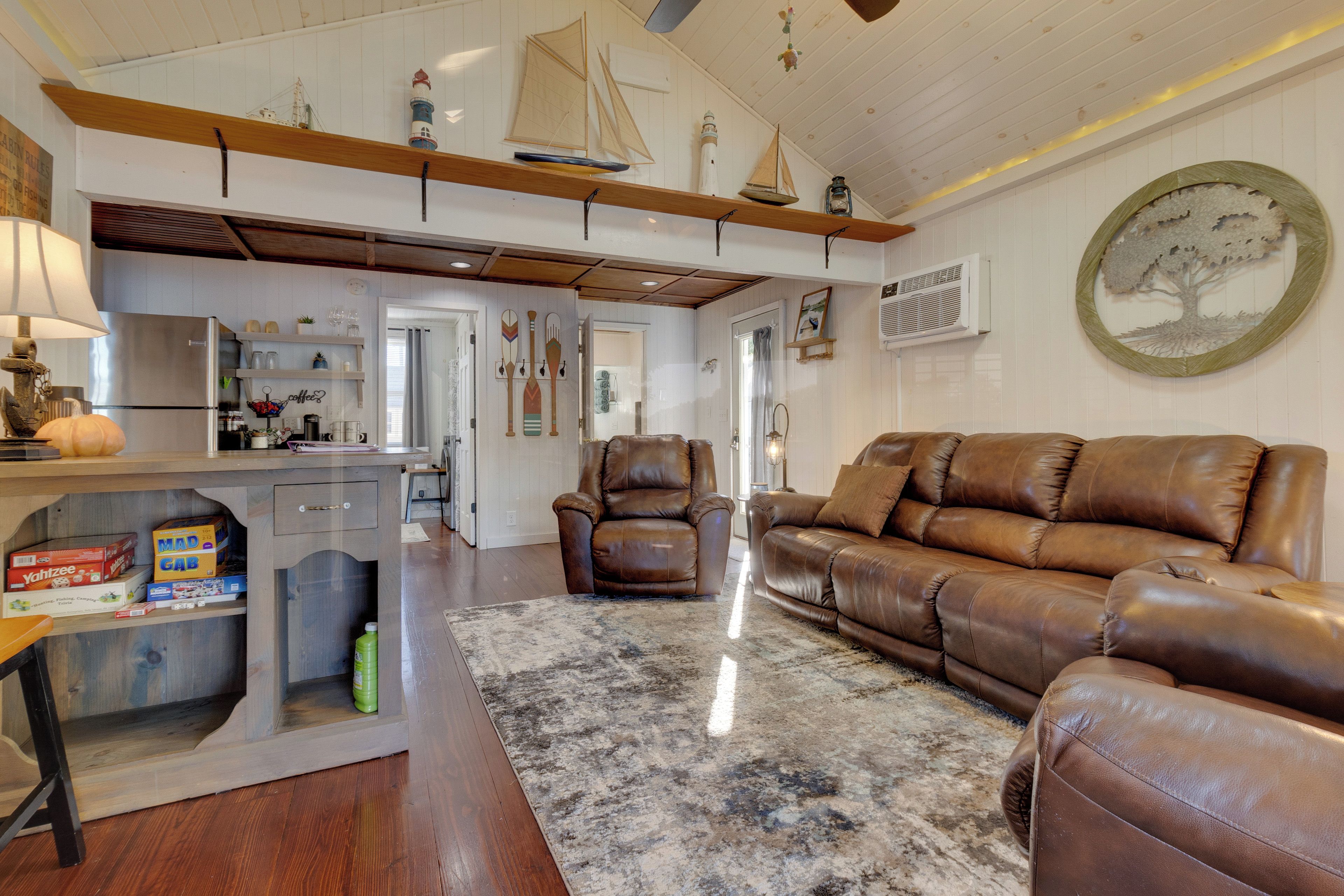 A cozy living room with a brown leather sofa, a coffee table, and a rug. There is a kitchen area with a refrigerator and a sink.