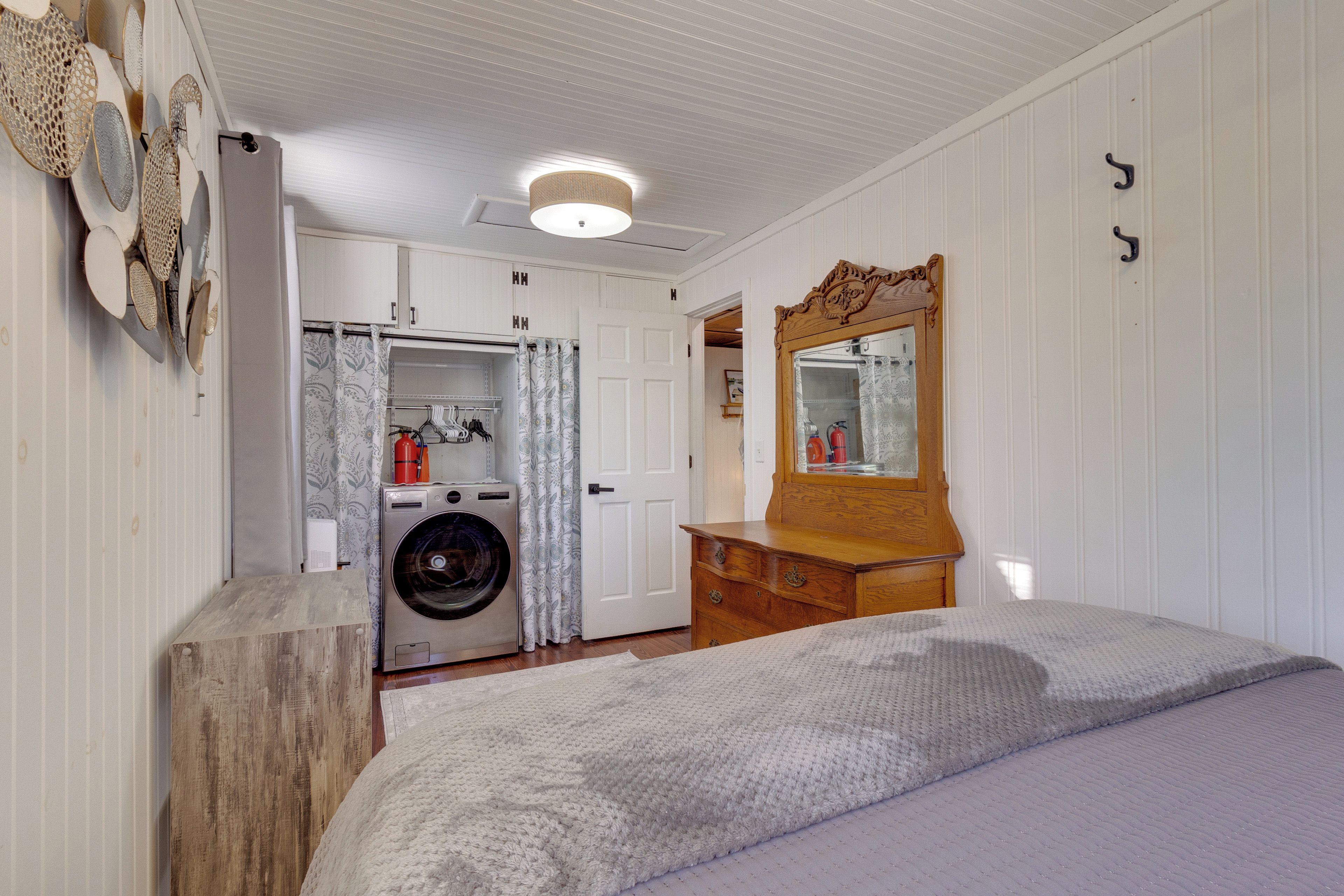 A bedroom with a bed, a wooden dresser, a mirror, a washing machine, and a door leading to a bathroom.