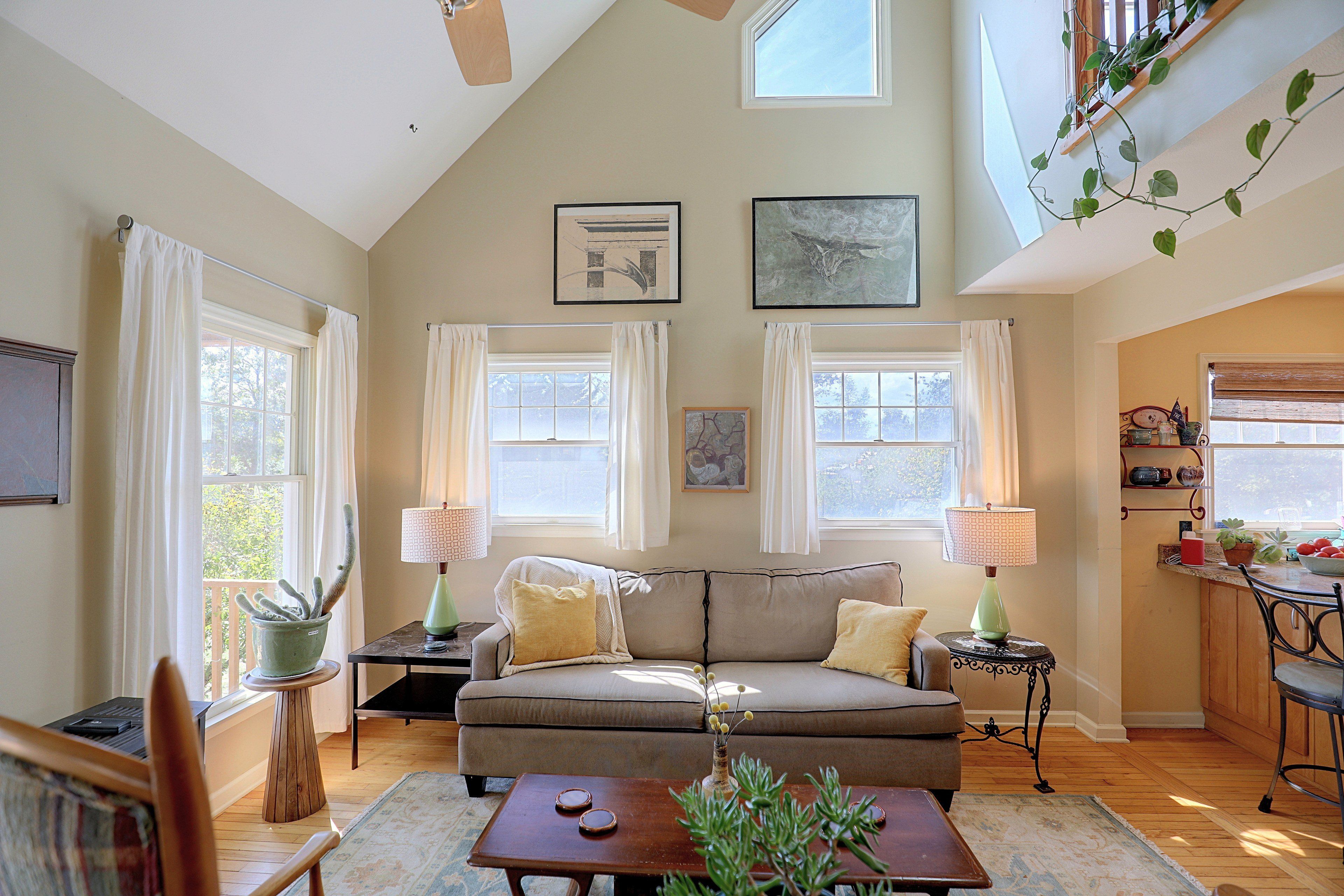 A living room with a sofa, two side tables with lamps, a coffee table, and a plant. There are windows with curtains, a ceiling fan, and artwork on the walls.