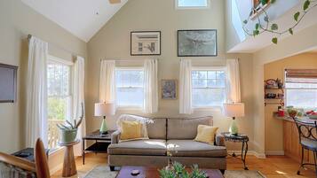 A living room with a sofa, two side tables with lamps, a coffee table, and a plant. There are windows with curtains, a ceiling fan, and artwork on the walls.