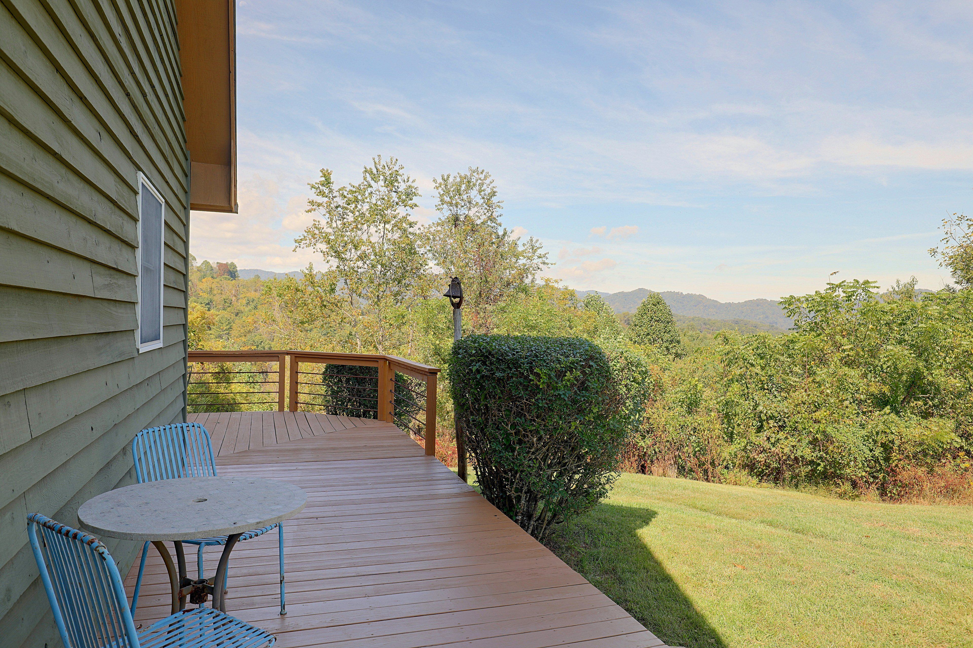 A wooden deck with a table and chairs, overlooking a grassy yard and trees.