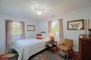 Un dormitorio con cama, una silla, una mesa y una lámpara. Hay cortinas en las ventanas y un candelabro cuelga del techo.