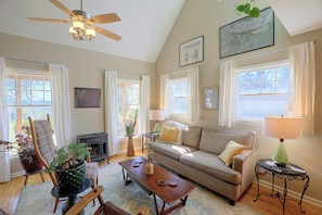 A living room with a sofa, coffee table, and a fireplace. There are windows with curtains, a ceiling fan, and artwork on the walls. - Misty Mountain, Peaceful Mountain Retreat (Marshall)