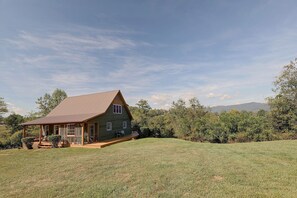 A single-story house with a sloped roof, a porch with seating, and a view of a grassy field and trees. - Misty Mountain, Peaceful Mountain Retreat (Marshall)