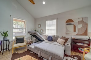 A room with a convertible sofa, two windows with blinds, a small table with a plant, and a wall with abstract art.