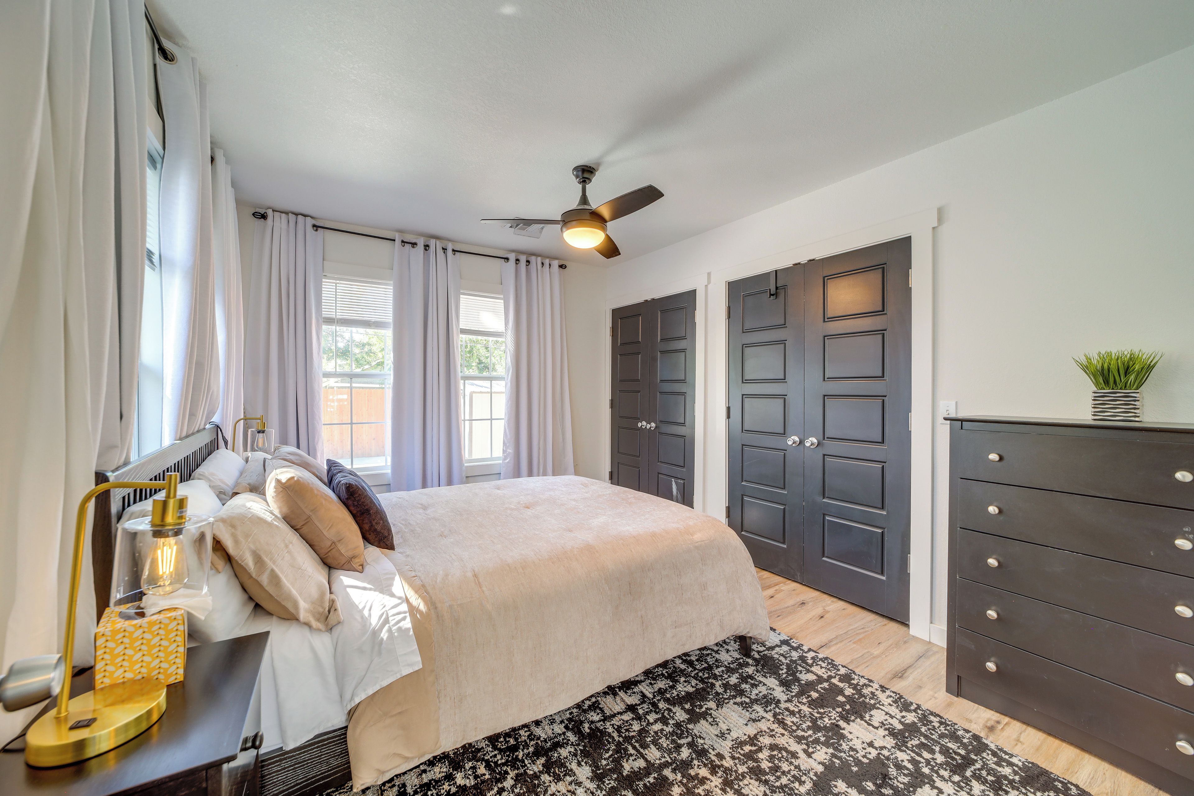 A bedroom with a bed, bedside table, lamp, curtains, windows, ceiling fan, and a dresser.