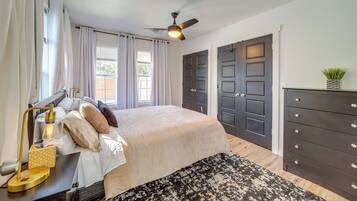 A bedroom with a bed, bedside table, lamp, curtains, windows, ceiling fan, and a dresser.