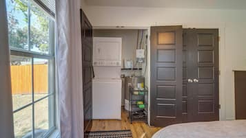 Een knus kamertje met een bed, een kleine kitchenette, een raam met gordijnen en een deur naar een andere kamer.