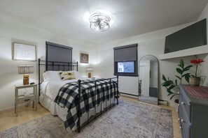 Een slaapkamer met een bed, nachtkastjes, een lamp, een raam met zonwering, een spiegel en een kamerplant.