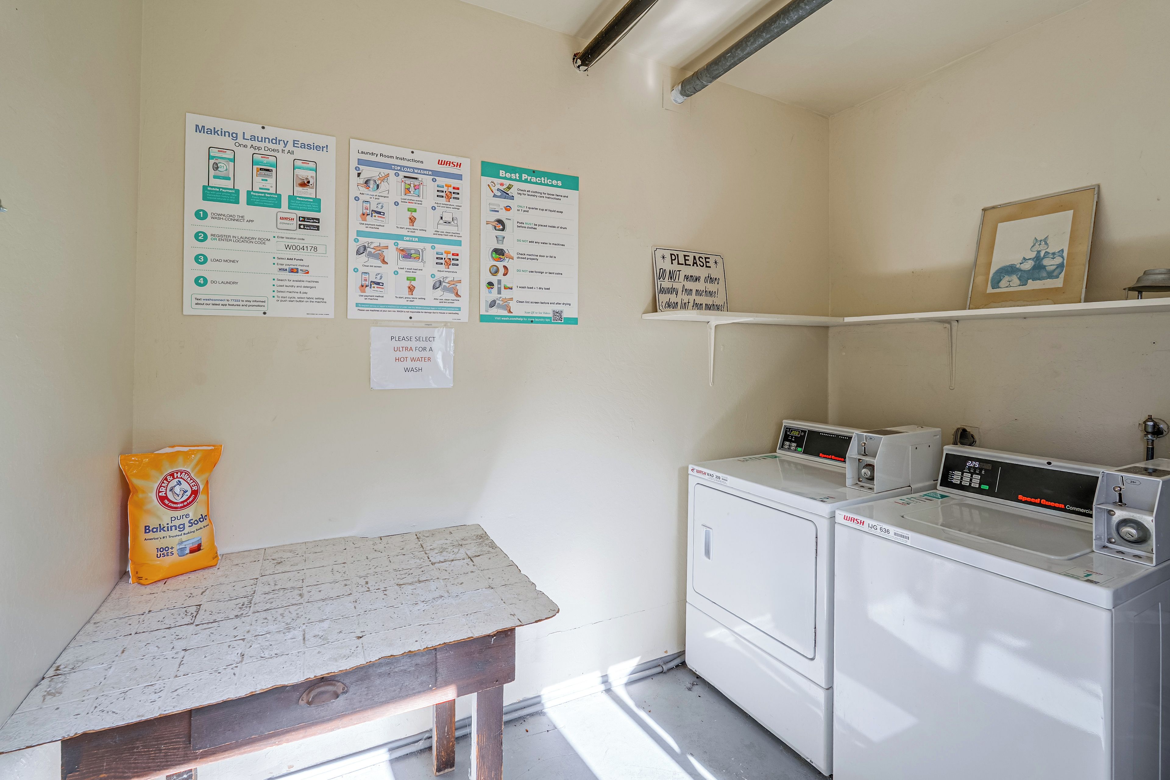 A laundry room with two washing machines, a drying machine, and a table with a bag of detergent. There are instructional posters on the wall.