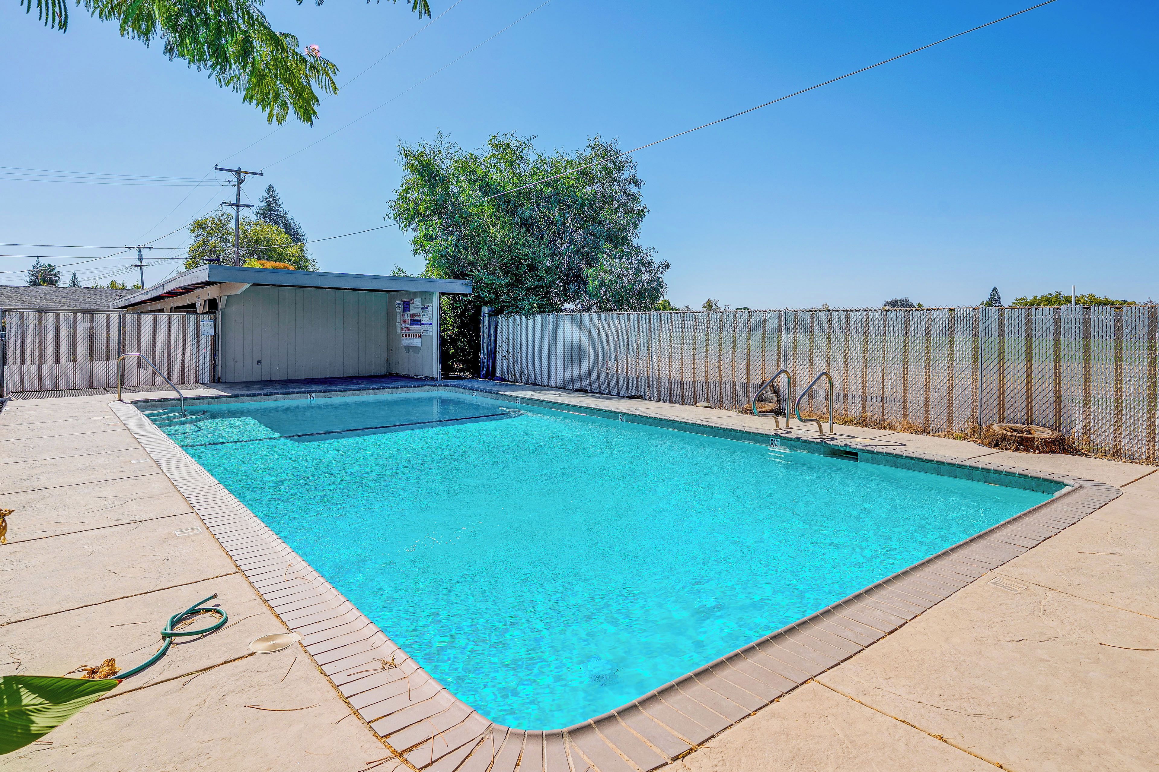 A rectangular swimming pool with a tiled deck surrounding it, a covered area with a door, and a chain-link fence enclosing the property.