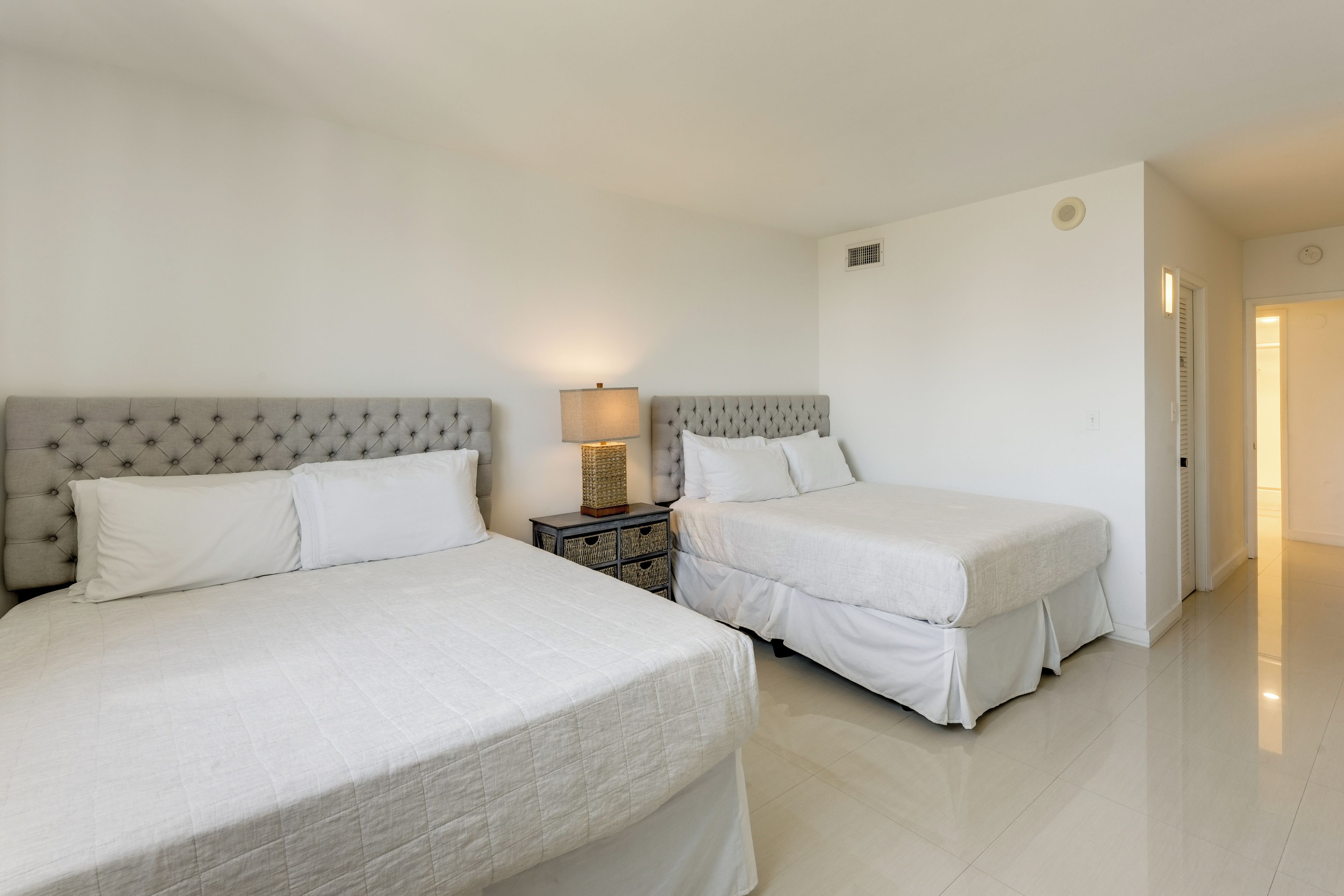 Una habitación de hotel con dos camas, una mesita de noche con lámpara y una puerta que da a otra habitación.