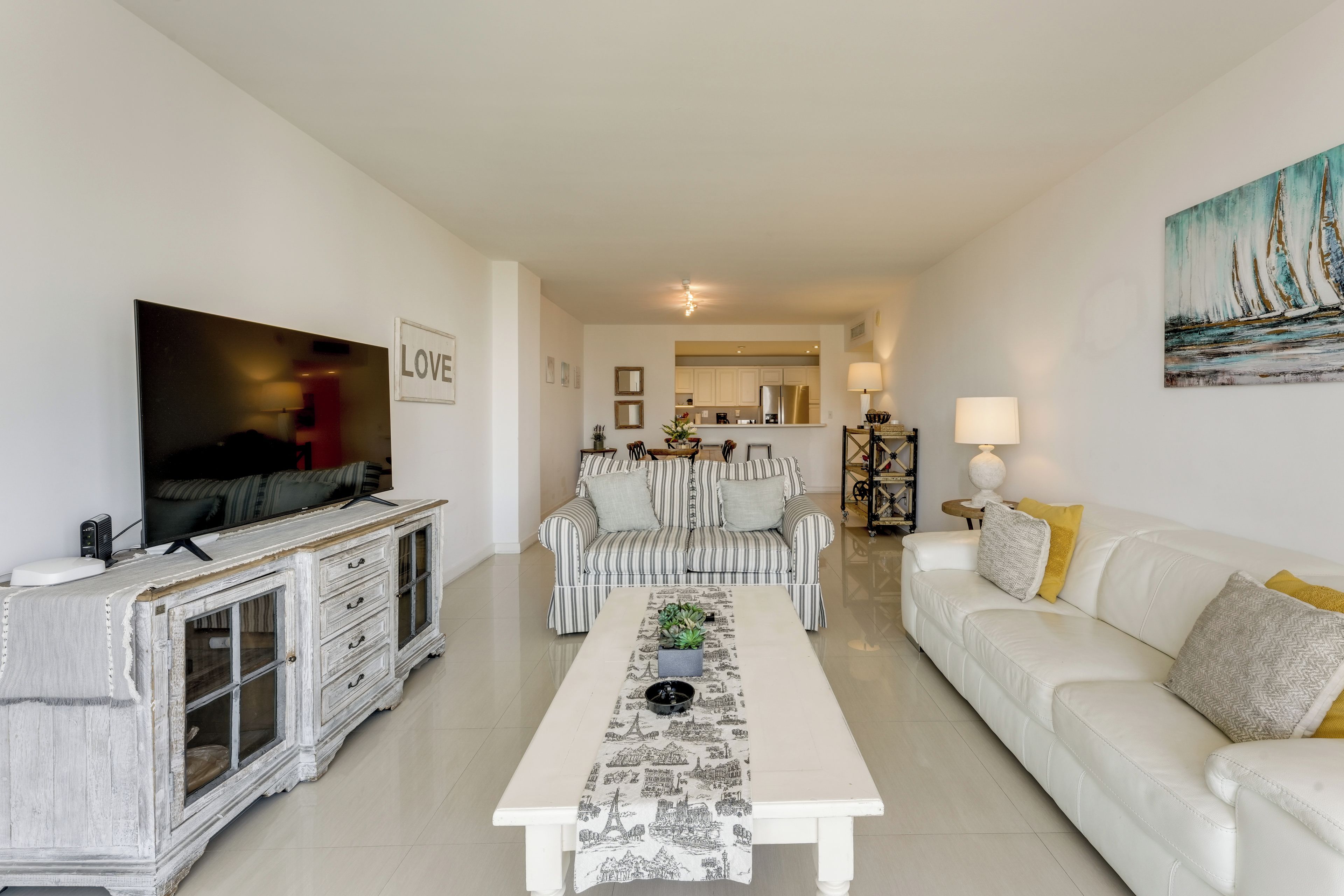 Una sala moderna con un televisor de pantalla plana, un sofá blanco, sillones a rayas, una mesa de centro con mantel de temática de viajes, y una cocina al fondo.