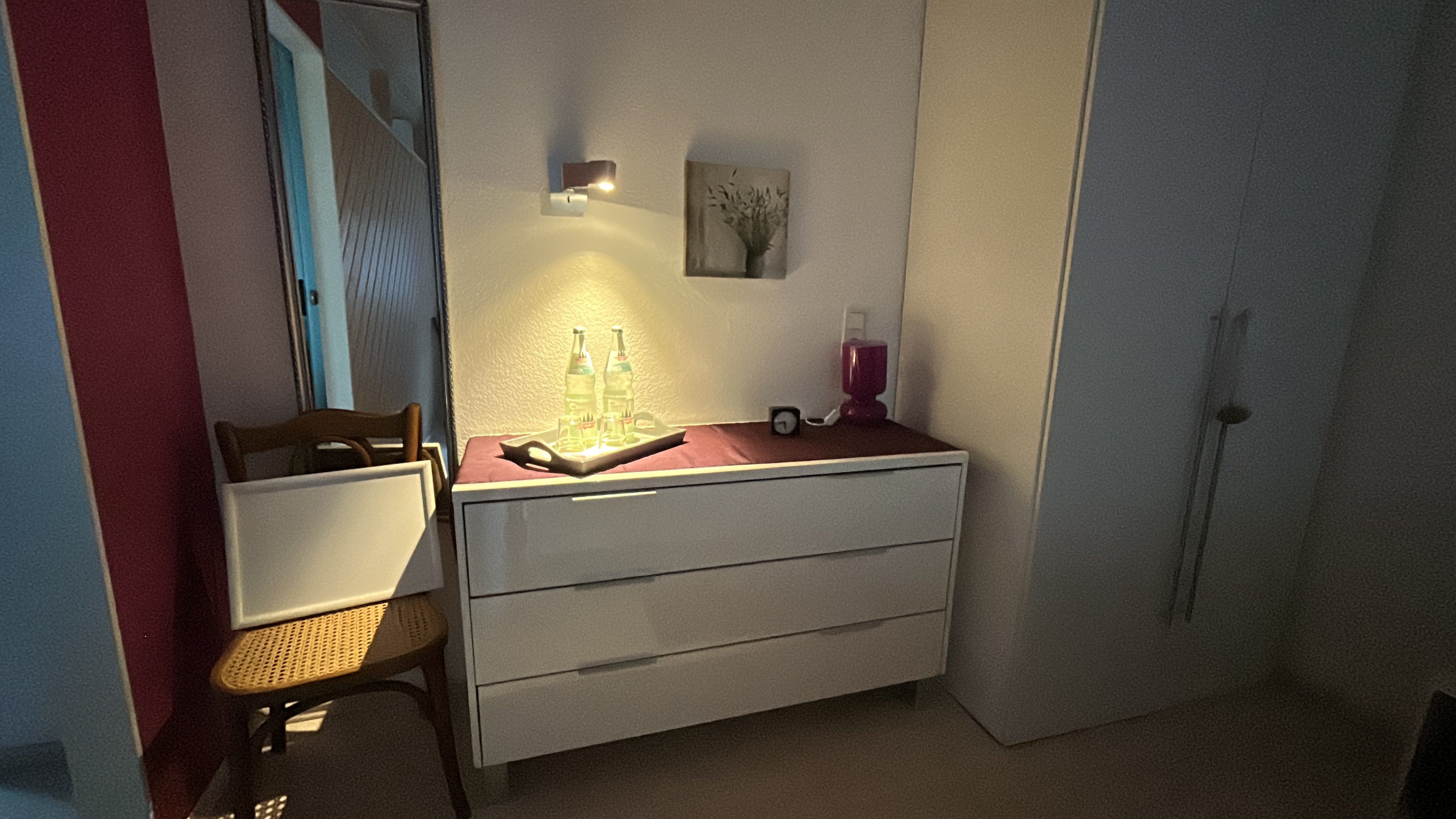 A small bedroom with a single bed, a bedside table with a lamp, a chair, and a wall-mounted shelf with bottles and a clock.