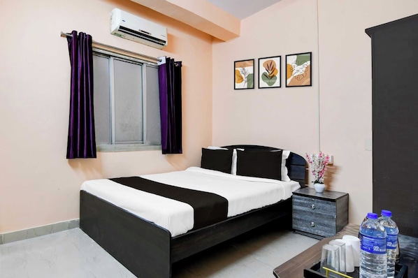A bedroom with a bed, a nightstand, a window with purple curtains, an air conditioner, and framed artwork on the wall.