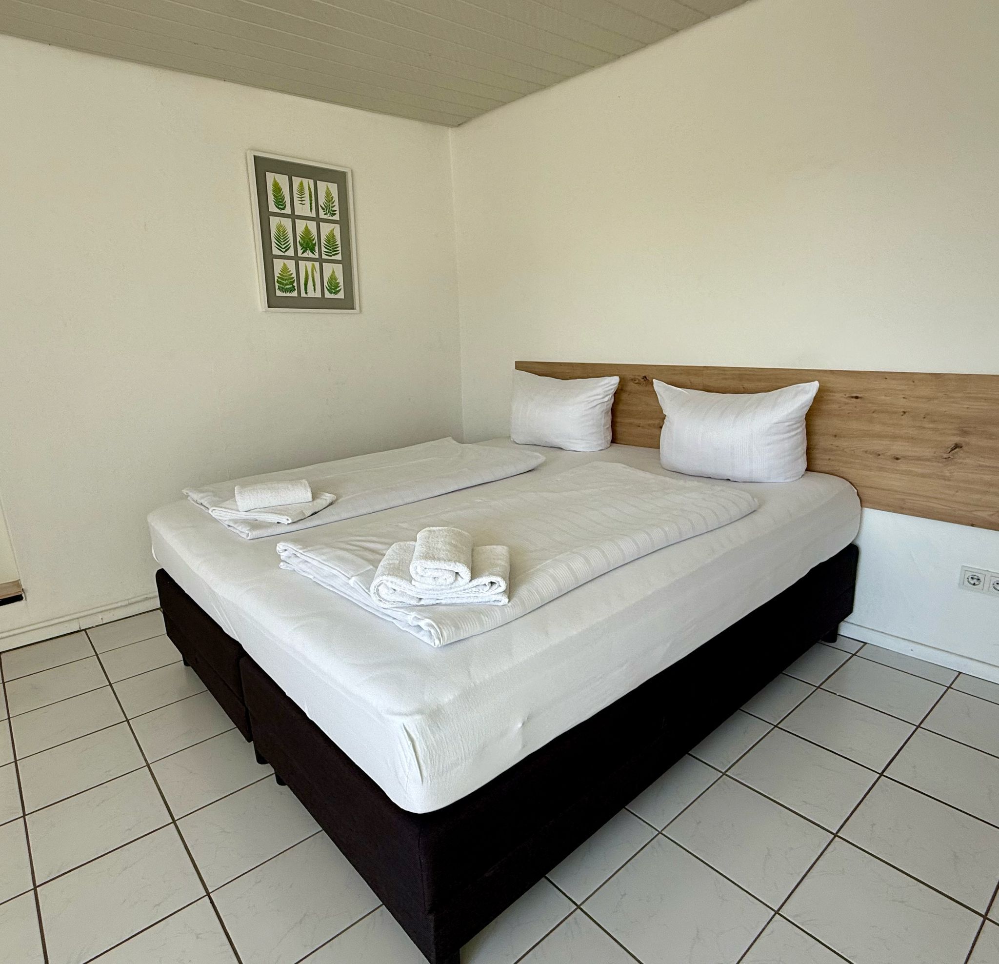 Quarto de hotel moderno e limpo, com uma cama grande, roupa de cama branca e um quadro com folhas na parede.