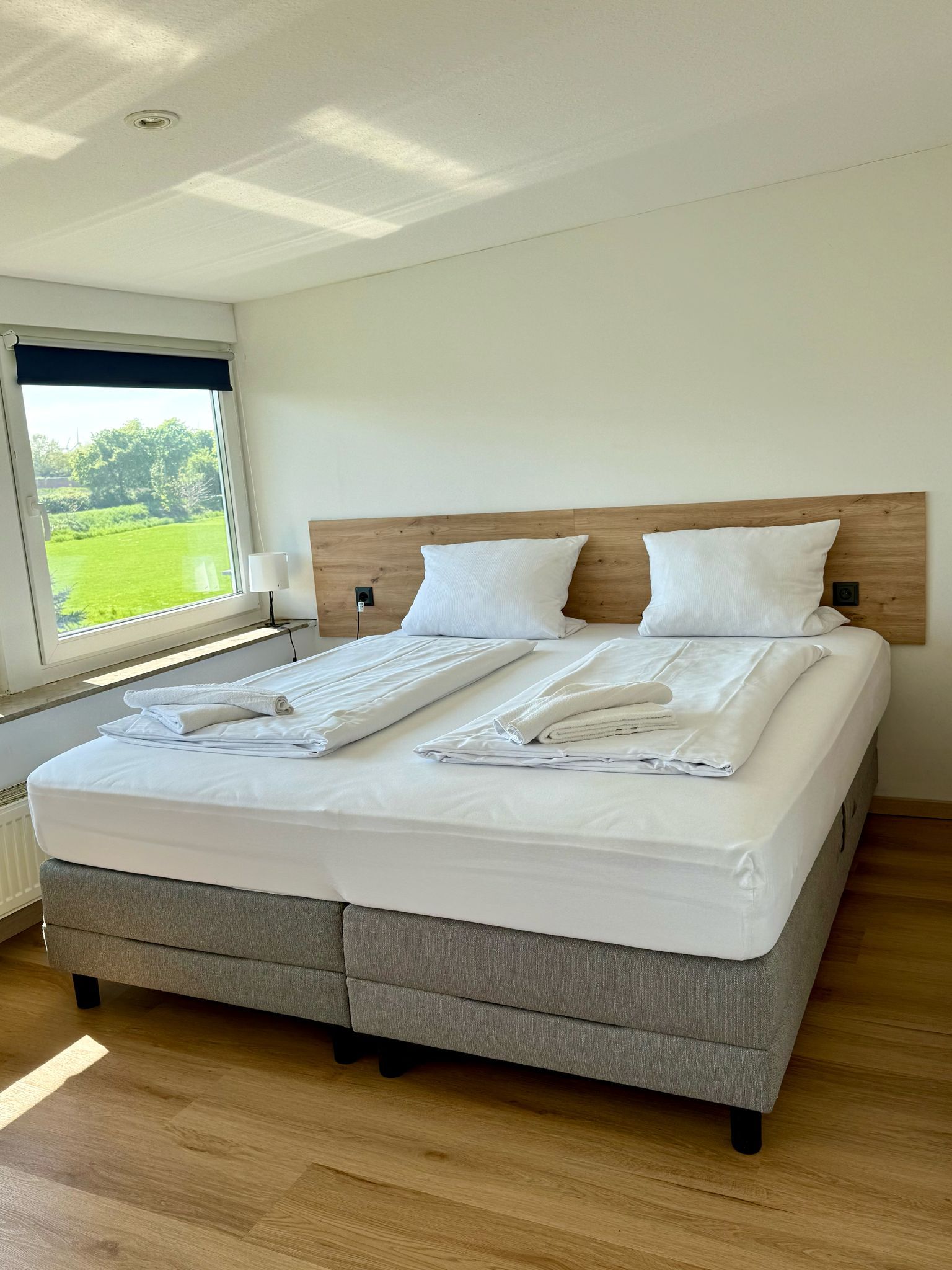 Ein ordentlich bezogenes Bett mit weißen Leinen und Handtüchern, ein hölzernes Kopfteil und ein Blick auf eine grüne Landschaft durch ein Fenster.