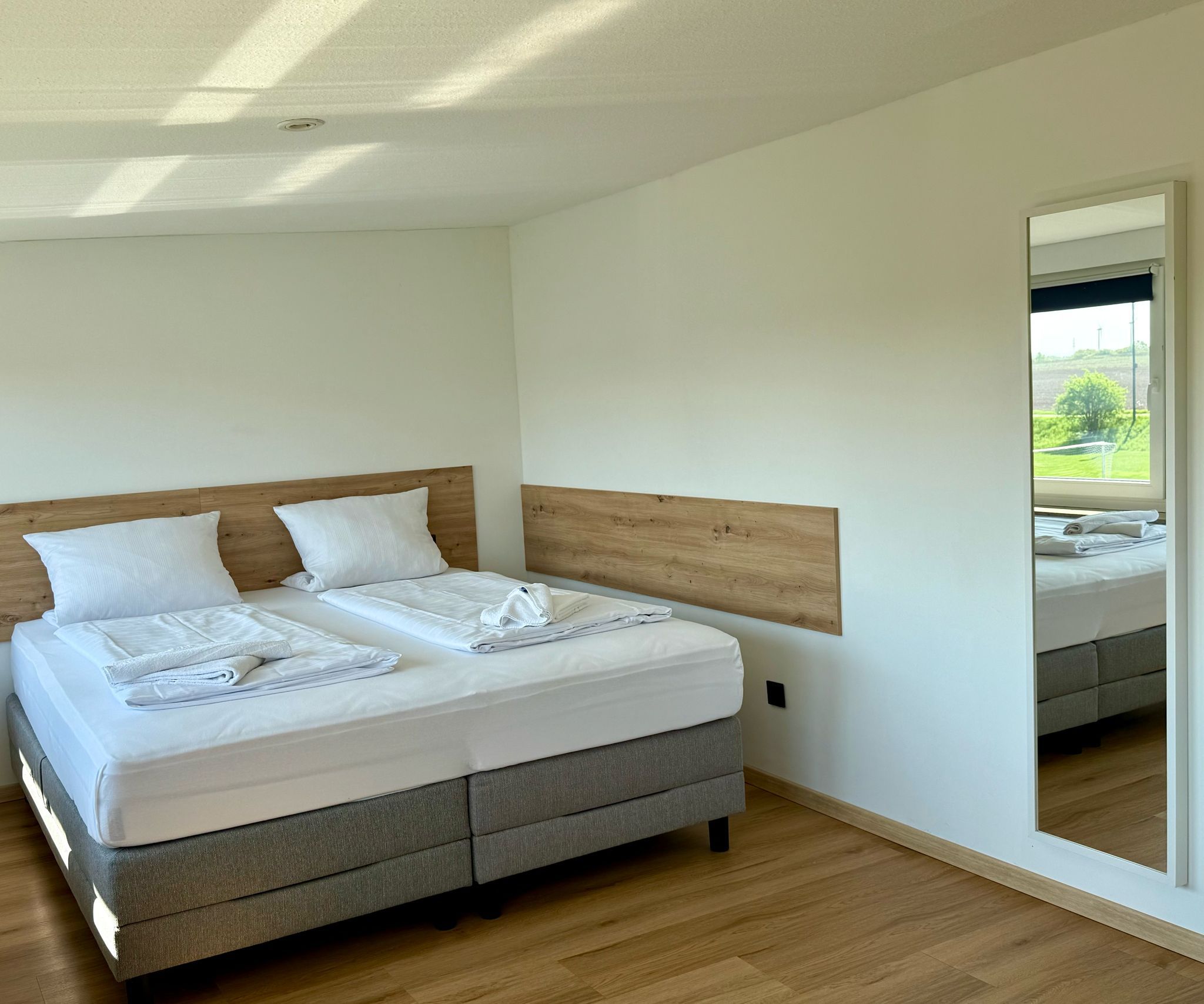 Une chambre moderne avec une tête de lit en bois, un grand lit recouvert de draps blancs soigneusement pliés, et une vue sur un paysage verdoyant par la fenêtre.
