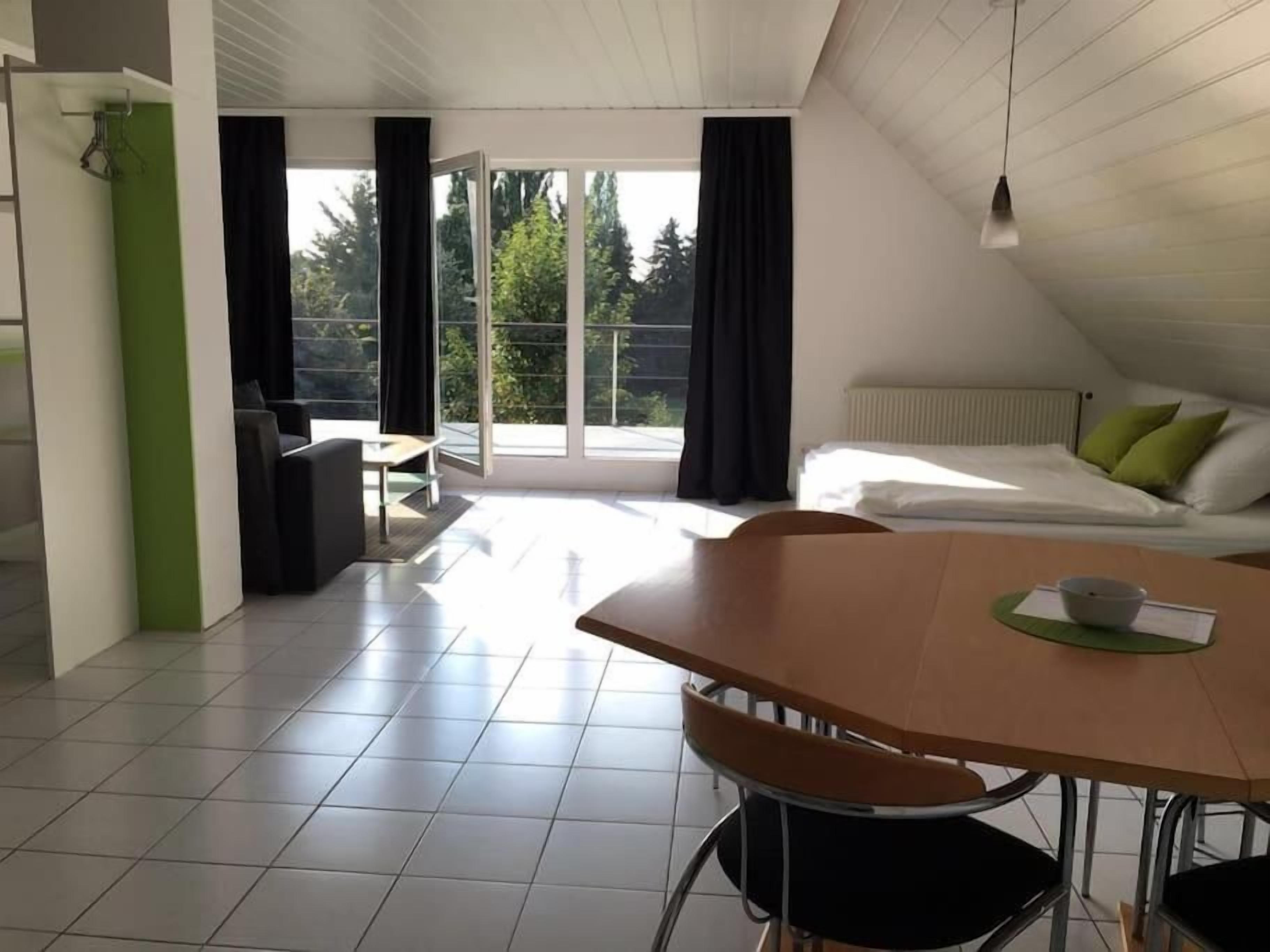 Ein modernes Zimmer mit Esstisch, Stühlen, einem Bett mit Kissen und Blick ins Freie durch ein großes Fenster.