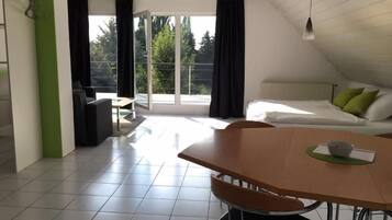Ein modernes Zimmer mit Esstisch, Stühlen, einem Bett mit Kissen und Blick ins Freie durch ein großes Fenster.