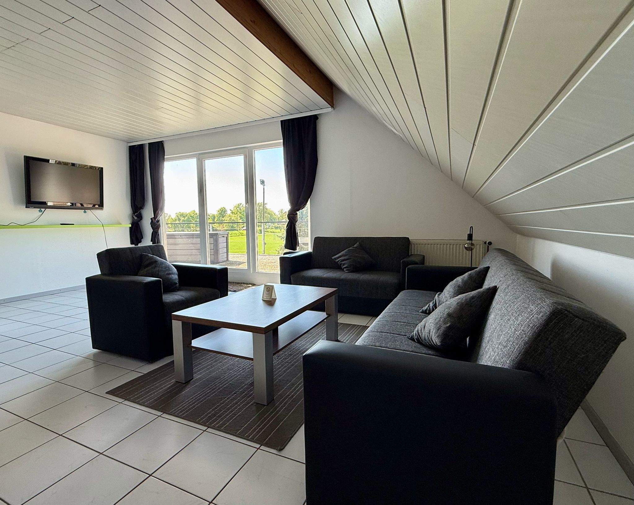 Ein modernes Wohnzimmer mit einem Flachbildfernseher, zwei Sofas, einem Couchtisch und einer großen Schiebeglastür, die auf einen Balkon führt.