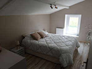 Ein Schlafzimmer mit Bett, Nachttisch, Lampe, Fenster, Heizkörper und Blick auf Grünflächen.
